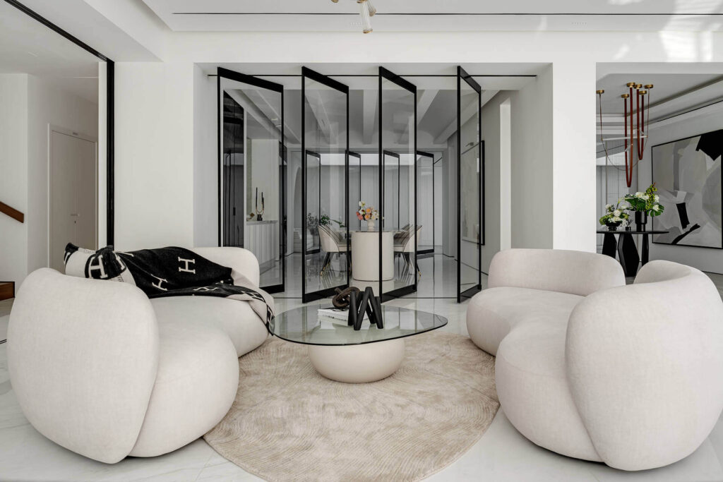 Modern living room with curved white sofas, round glass coffee table, beige rug, and black-framed folding glass doors.